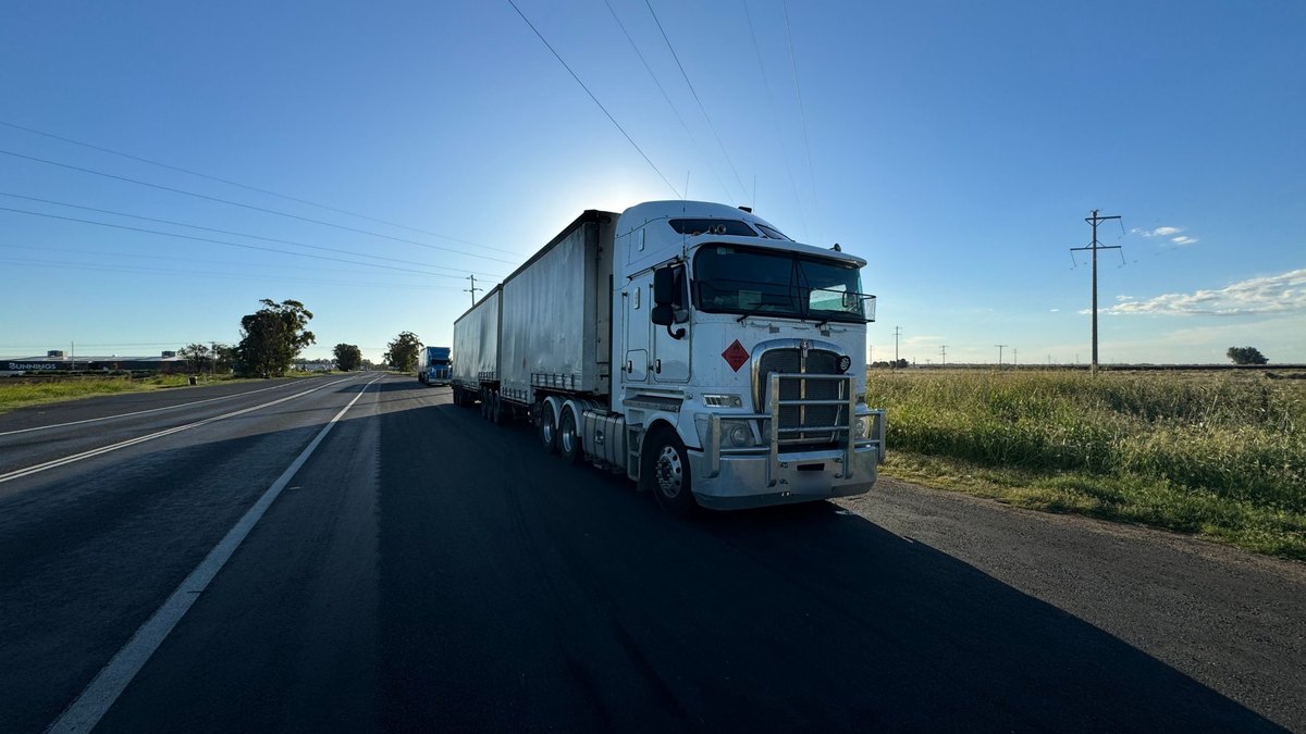 A white Kenworth K200 B-double parked on the shoulder of a flat country highway, low sun behind the cab