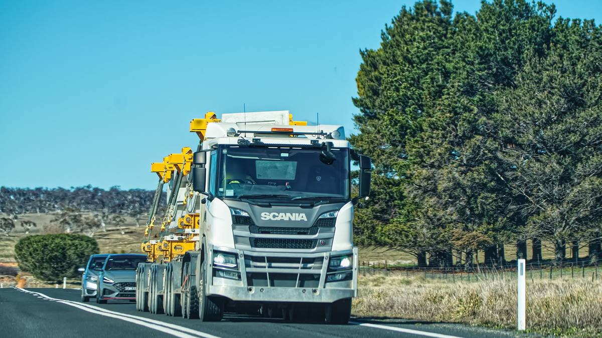 A Scania prime mover carrying a yellow oversize load on a rural road, with a car following behind