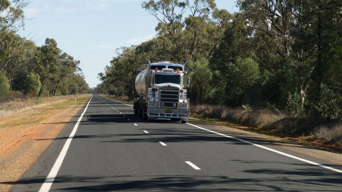 A Kenworth prime mover hauling a stainless tanker along a straight two-lane regional highway lined with gum trees